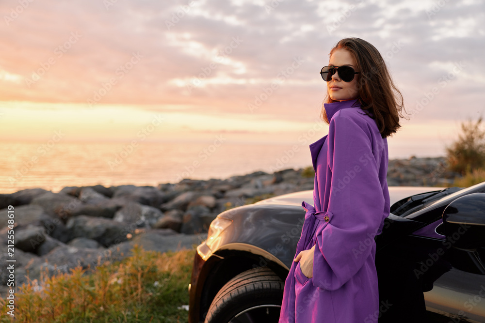 Fototapeta premium Woman lifestyle scene at seaside during sunset, wearing a purple coat and sunglasses, leaning beside a sleek car, captures confident pose and relaxed mood in golden hour light