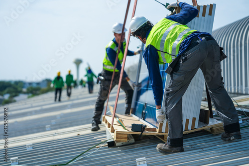 Bild auf Leinwand A solar installation engineer in a white helmet and high visibility vest carefully bends over the pallet checking each module label on the curved metal roof of an industrial factory