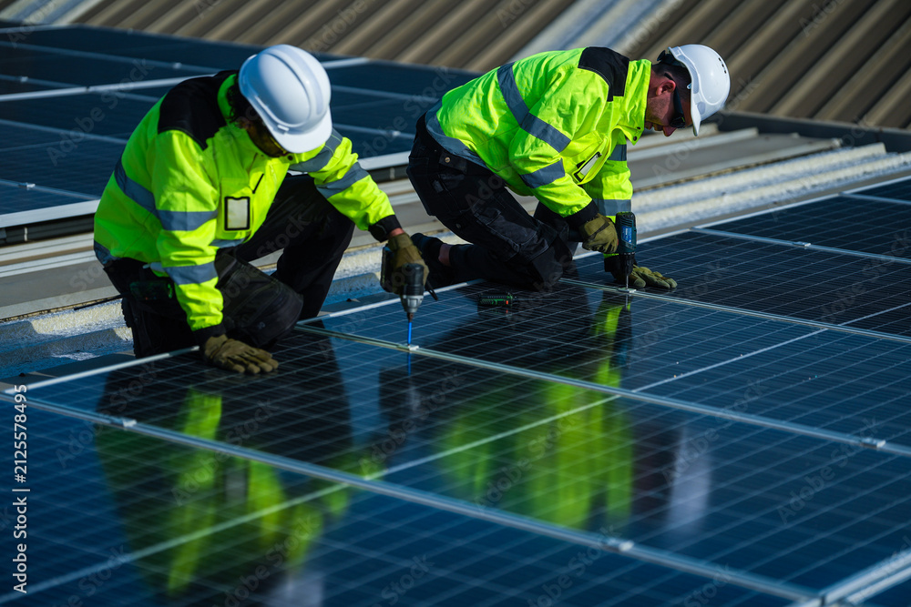 Fototapeta premium A solar installation engineer in a white helmet and high visibility vest confidently kneels on the evening array reporting over the radio on a commercial factory rooftop photovoltaic.