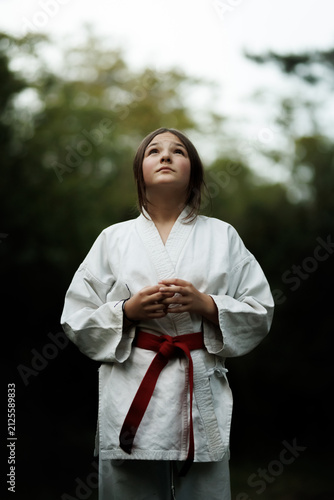 Canvas Print Young girl in karate gi with red belt standing outdoors looking up with calm foc