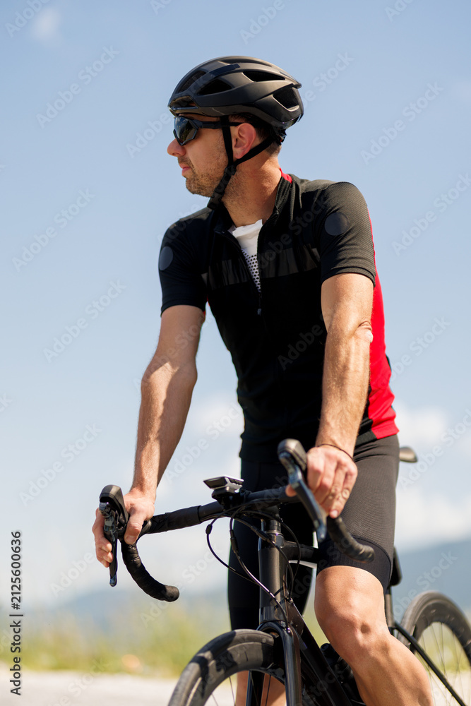 Fototapeta premium Cyclist in a red and black jersey takes a break on a sunny day while resting on a road bike at a scenic location