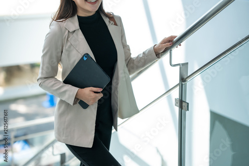 Smiling businesswoman in a beige blazer walking up stairs indoors while holding a tablet and using the handrail in a modern office building.