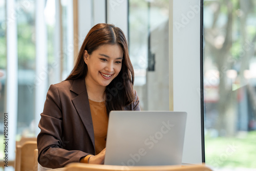 Smiling young woman using a laptop in a bright office, working at a table near a window.