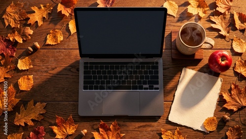 A laptop and mug of drink sit on a rustic wooden table surrounded by colorful autumn leaves.