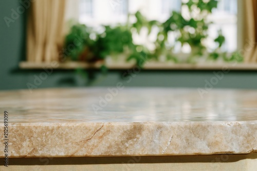 Polished tan marble table top with a honed edge and natural veining for product display against a soft blurred kitchen window with green foliage