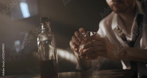 Close-up of a seated man holding a glass of whiskey in both hands beside a bottle on the table.