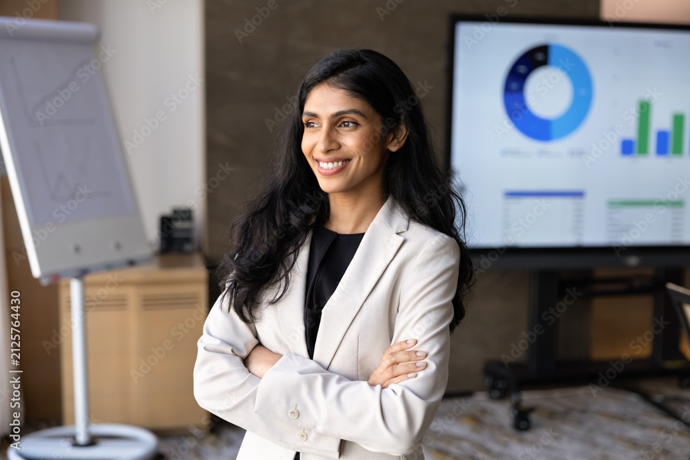 Obraz premium Portrait of Indian businesswoman or corporate trainer standing with crossed arms in corporate office during business presentation, financial charts and graphs displayed on digital screen in background