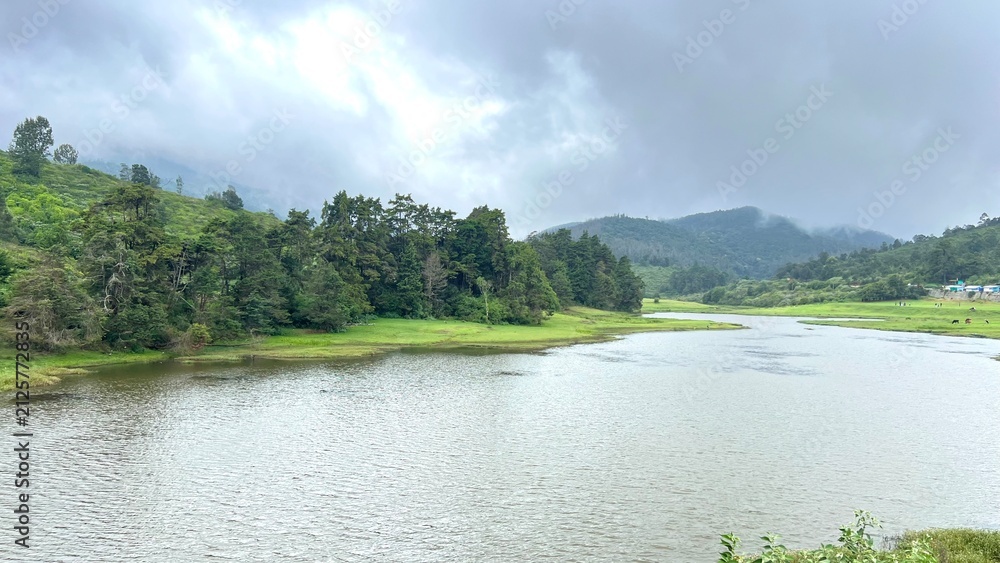 Obraz premium Scenic mountain lake surrounded by lush green hills, forest trees and misty clouds under overcast sky, capturing peaceful monsoon landscape, nature travel and hill station countryside view.