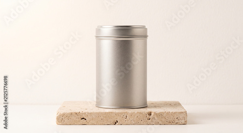 A sleek silver metal tin container with a lid standing upright on a textured beige stone block against a light background with copy space