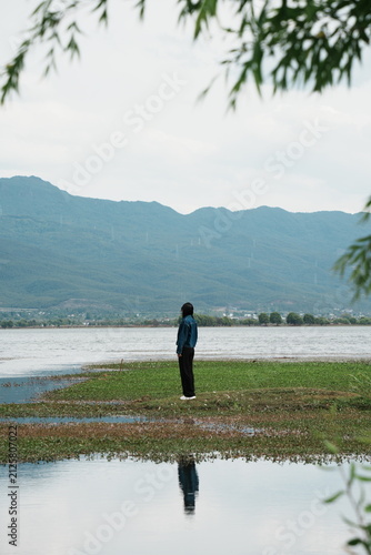 Foto Back view of lonely young woman standing on lakeshore, calm water reflection and