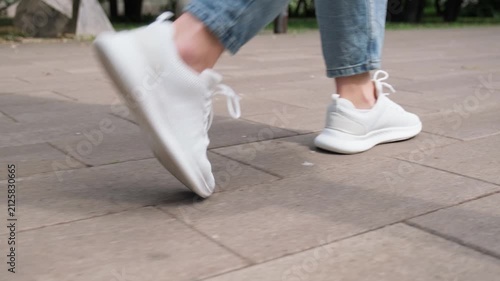 Person strolling outdoors in comfortable white footwear. Low angle shot of a person wearing casual clothes and white running sneakers walking on a city sidewalk. Casual active lifestyle concept.
