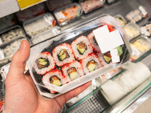 A shopper holding a package of sushi in a store. A hand holds a plastic box with sushi rolls against a supermarket shelf, symbolizing ready-to-eat food and convenience