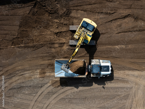 Foto Aerial view of a yellow excavator loading soil into a white dump truck at a construction site with visible tire tracks in the dirt in Kaunas, Kaunas County, Lithuania