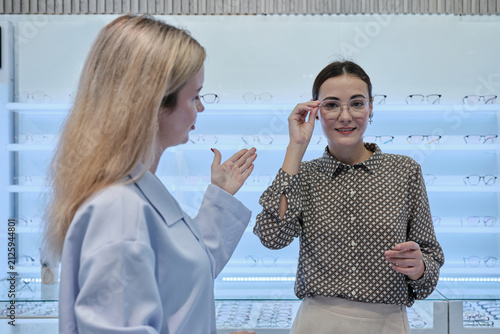White female customer chooses eyeglasses with optometrist's help and guidance, improved eyesight for individual lifestyles, consults on decisions, and options choices at store display in optical shop.