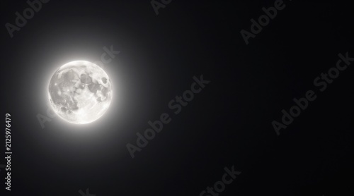 Full moon shines brightly with a glowing halo against a dark night sky background. Glowing full moon illuminates the dark sky with a soft radiant halo surrounding it.