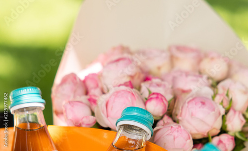 Close up of outdoor event details with floral arrangements with roses, lisianthus, and anemones, wrapped bouquets, and beverage bottles placed on a table against a blurred garden background.
