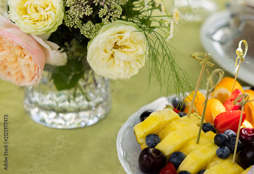 Close up of outdoor event catering featuring sliced cured meats, toasted bread with pesto, assorted fruit platters, and cheese boards. The appetizers are arranged on a table in natural daylight.