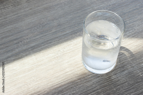 Glass of cold water with ice cubes under natural sunlight casting soft shadow on wooden table background.