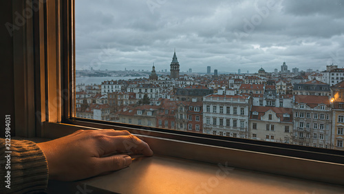Close-up of a person hand resting on a windowsill, looking out at a grey overcast autumn sky over an unfamiliar foreign city skyline. Adjustment and distance from home concept.
