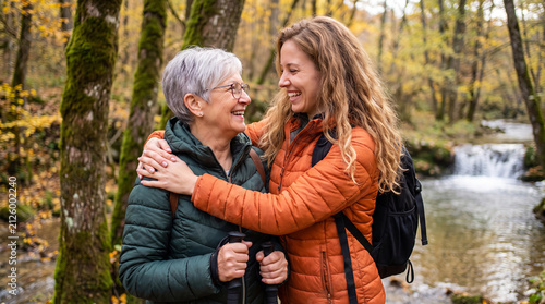 Two women embrace while hiking in a forest, surrounded by autumn foliage and a flowing stream in the background