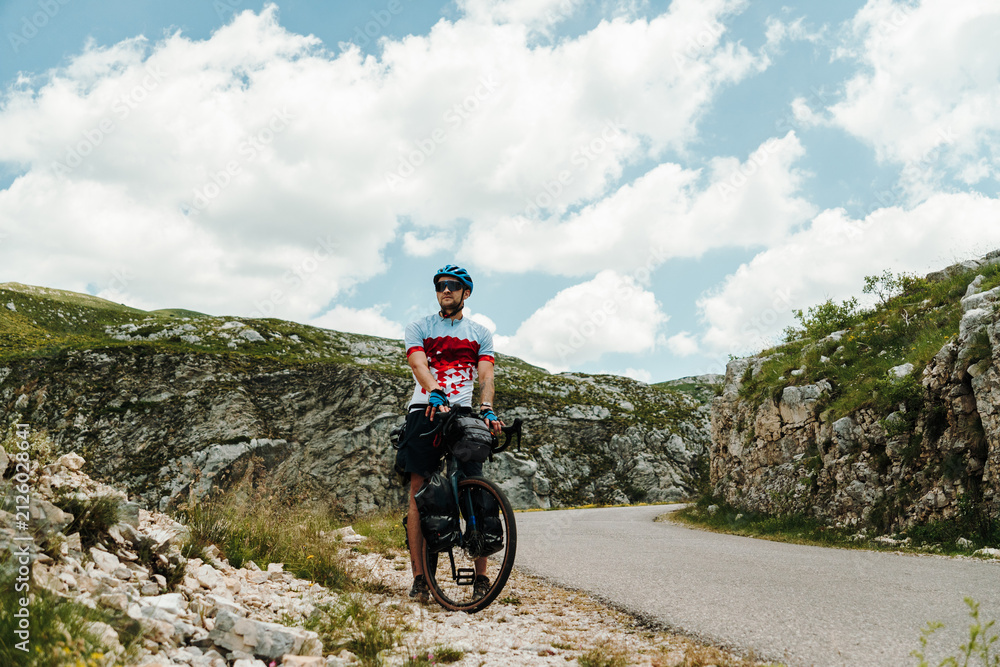 Fototapeta premium Bikepacking cyclist pauses beside a winding mountain road in Durmitor National Park, Montenegro. Loaded gravel bike and rugged alpine scenery capture the spirit of adventure travel