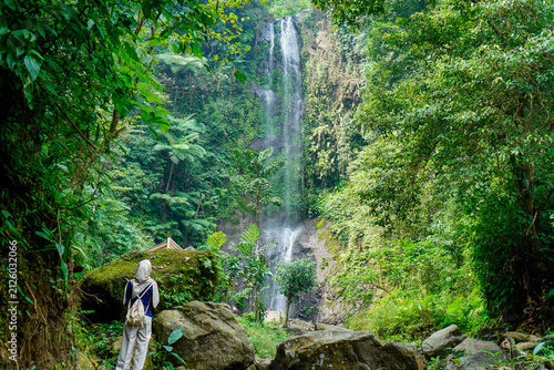 ​A female traveler wearing a hijab and carrying a backpack stands on a rocky path, looking up at a magnificent tall cascading waterfall surrounded by dense, lush green tropical rainforest foliage. Con