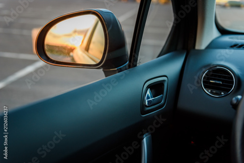 Left side rearview mirror inside a car. Close-up view of the vehicle's exterior left mirror and interior driver side panel during sunset.