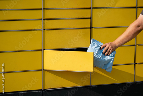 Hand placing blue package into yellow delivery locker with multiple compartments visible