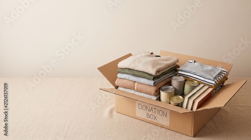 cardboard donation box filled with clothes, canned food, and books, soft natural window lighting, warm neutral color palette, clean empty space in the upper half for text