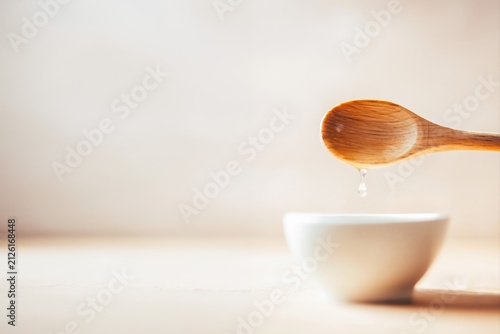 A wooden spoon with a droplet falling into a white bowl on a light background. Minimalist composition highlights natural kitchen utensils and clean food preparation.