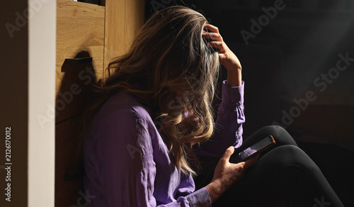 Depressed woman holding smartphone sitting on floor in darkness.