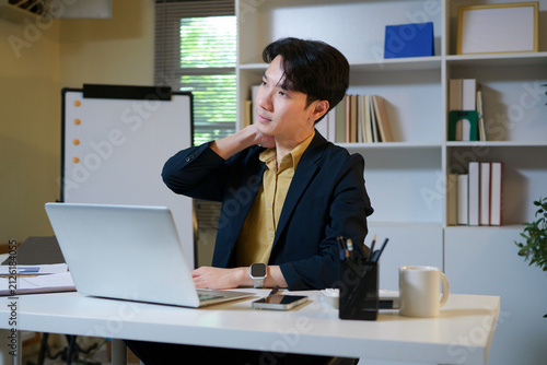 Asian businessman suffering from acute neck pain and discomfort, touching his sore neck while working on a laptop at his desk in a contemporary office setting, experiencing stress and health issues