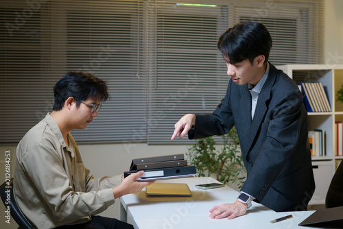 Quadro su tela an Asian male supervisor or manager, wearing a business suit, stands leaning over his desk and pointing his finger, giving orders or reprimanding male employees