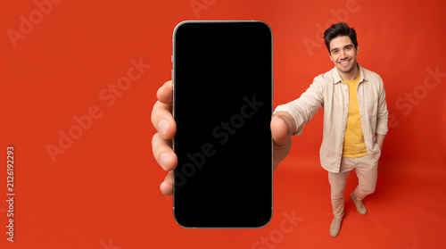 Smiling young man presenting blank black smartphone screen towards camera on vibrant red background for app mockup