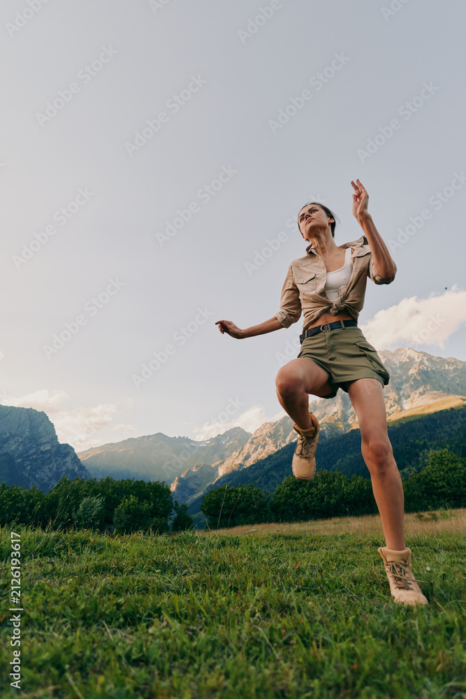 Obraz premium Woman jumping in a meadow with mountains in the background, casual outdoor attire, bright sky, and grass underfoot, capturing a vibrant lifestyle and energetic outdoor moment.