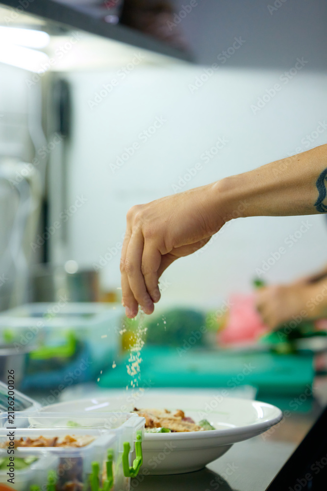 Fototapeta premium Chef seasoning fresh salad during plate assembly in commercial kitchen. Concept of restaurant plating, fresh meal preparation, professional food service.