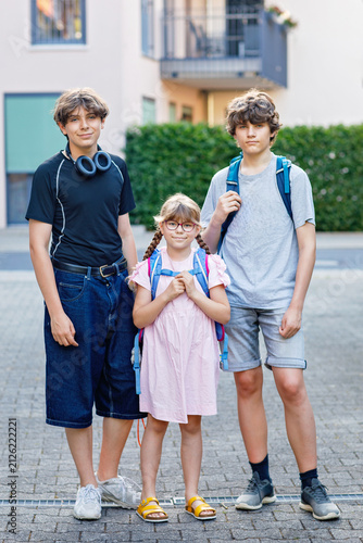 Three siblings with backpacks standing together outdoors before school in a residential neighborhood. Two teenager boys and little girl. Brothers and sister.