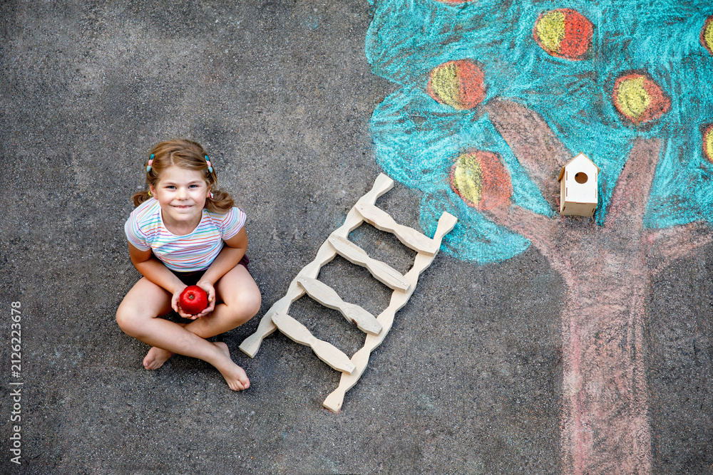 Naklejka premium Cute little girl painting with colorful chalks apples harvest from apple tree on asphalt. Cute preschool child with having fun with chalk picture. Creative leisure for children, drawing and painting.