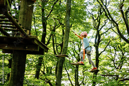 Happy little girl climbing rope course in forest adventure park, child wearing helmet and harness enjoying outdoor activity, real life moment. School child having fun.