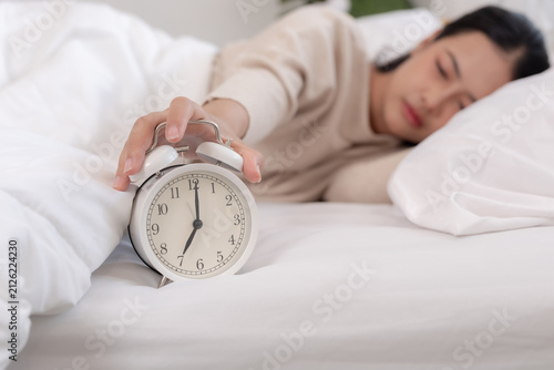 Tired Asian woman in comfortable bed reaching hand to snooze white alarm clock during early morning showing sleepy reluctant expression about waking up inside cozy bedroom.