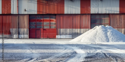 Potash fertilizer pile in front of a large industrial warehouse with red and gray corrugated metal siding and large doors, showcasing a manufacturing environment