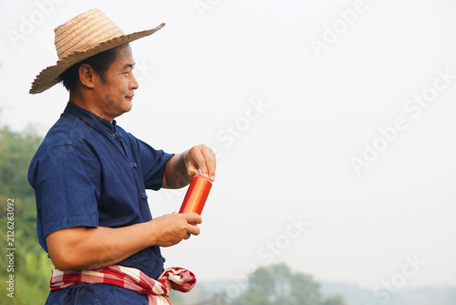 Asian farmer wearing a straw hat and traditional blue shirt holding a beverage can outdoors. Rural lifestyle, agricultural work, relaxation, and countryside living with copy space for advertising.