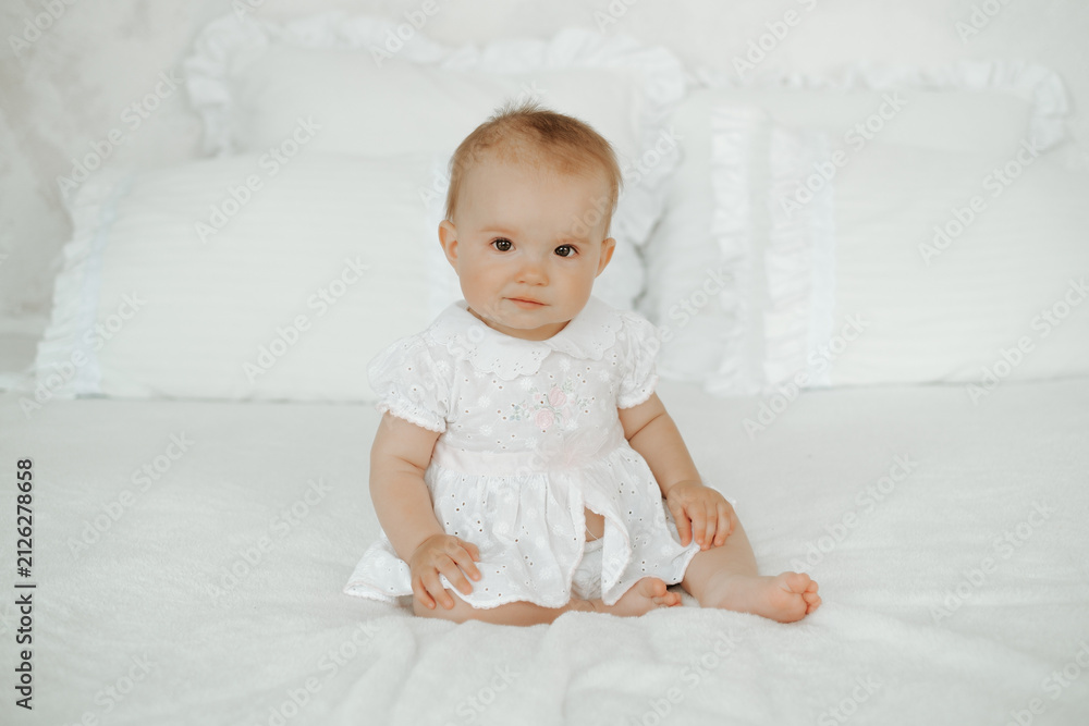 Fototapeta premium A happy baby is on the bed at home; a smiling little girl is sitting on a white cotton bed before going to sleep; a happy little kid, space for text.