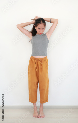 Portrait of cute young Asian girl tying her hair into a ponytail while standing and smiling at camera, stands barefoot against white wall room.