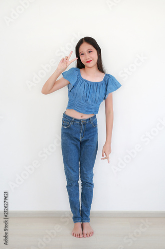 A young Asian girl in denim crop top and jeans standing barefoot, giving the peace sign, indoors against white background.