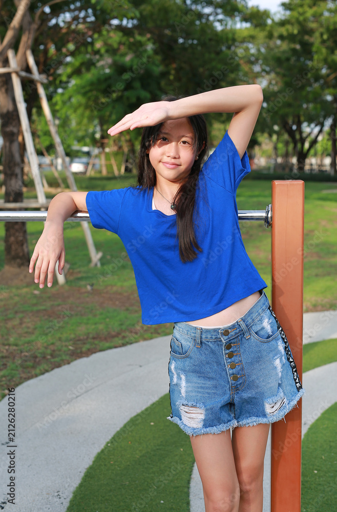 Obraz premium A young girl in a blue shirt is leaning against the playground bars, shading her eyes with her hand from the strong sunlight.