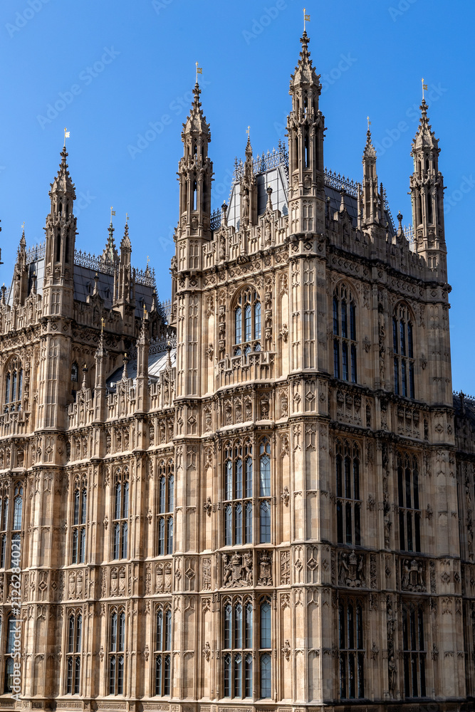 Obraz premium Detailed view of the ornate Gothic Revival architecture of the Palace of Westminster in London, England, showcasing intricate stonework, towers, and historic windows beneath a clear blue sky