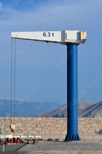 A vertical shot of a blue and white harbor jib crane with a 6.3 tons capacity label standing on a stone pier with mountains in the background.