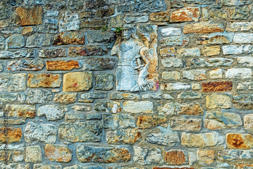Weathered carved stone figure incorporated into an old masonry wall, surrounded by irregular natural stone blocks. The historic architectural detail shows visible signs of age and erosion.