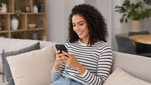 Happy young woman with curly hair using smartphone on sofa. Smiling female texting on mobile phone in living room